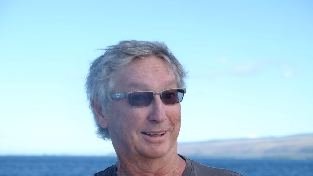 headshot of male sporting sunglasses and with a head of white and brown hair and with the ocean behind him.