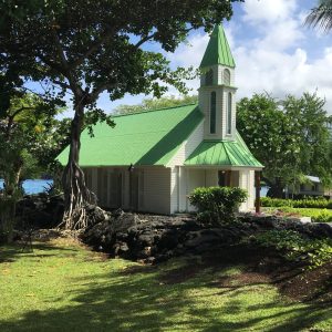 Classic Hawaiian style small wooden white church with green roof surrounded by walls of black lava, ohia trees and the Pacific Ocean in South Kona