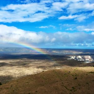 Halemaumau caldera on Kilauea Mountain with active outgassing in white to the right and a rainbow to the left and with puffy white clouds and blue sky overhead