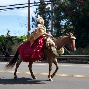 Hawaiian Princess on horseback wearing a gold and red dress and adorned with flowers in the Kamehameha Day parade in Hawi