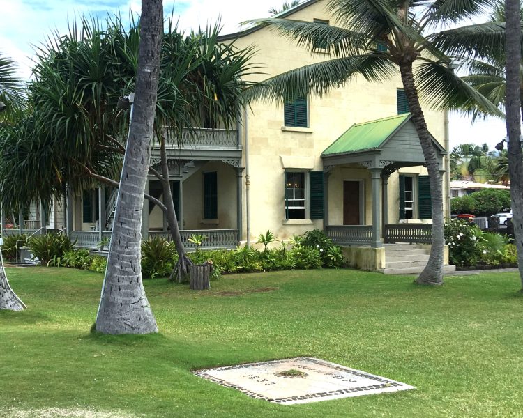 The Hulihee Palace in Kailua, Kona along the shore with palm tree fronting the yellow, green, and gray 2 story storied palace